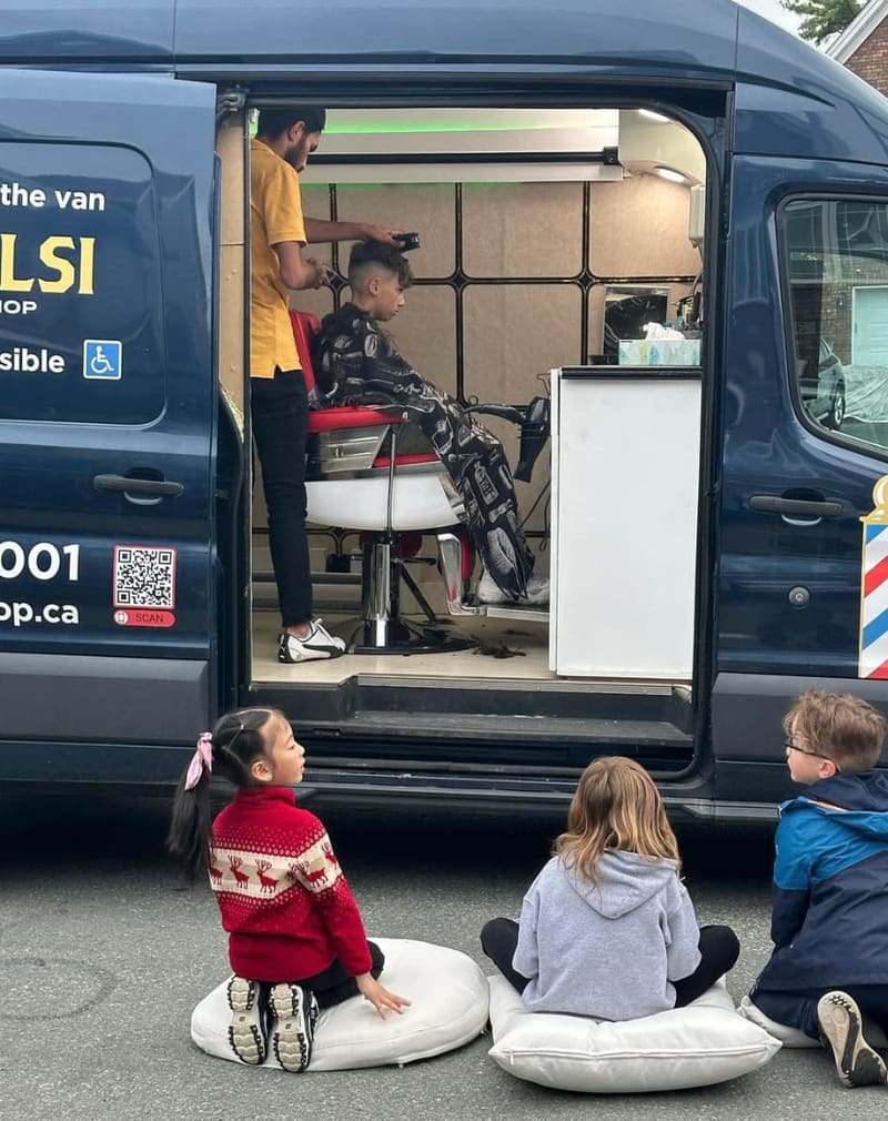 The side door of the van open on a haircut in progress, three kids sitting on cushions outside watching